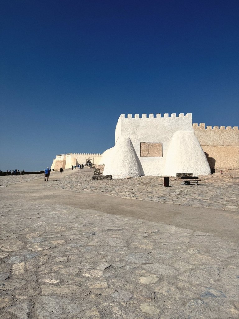 Whitewashed buttresses and crenellated walls of the hilltop kasbah, with visitors walking up the cobbled approach to the gateway under a deep blue sky