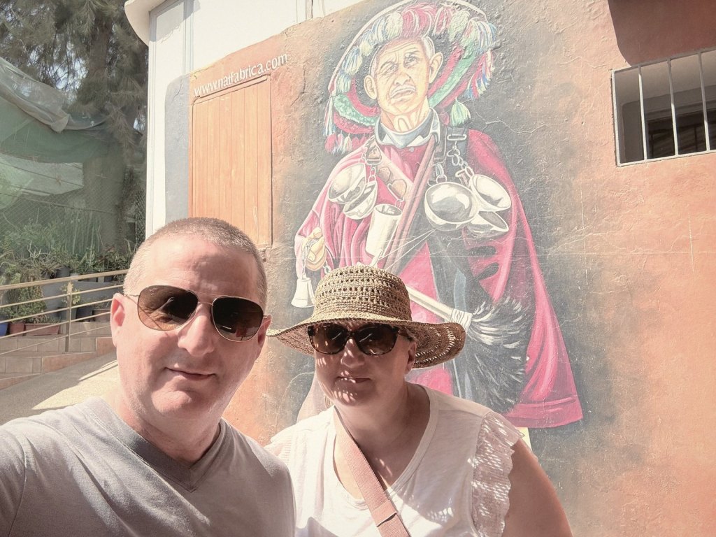 Chris and Jen, in sunglasses, taking a selfie in front of a large mural of a Moroccan water seller in a red robe and wide fringed hat hung with brass cups