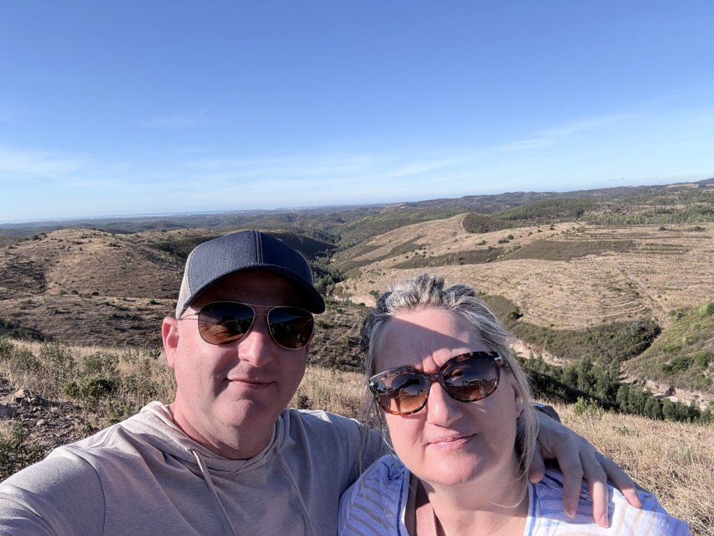 Chris and Jen in sunglasses taking a selfie above rolling dry hills, with the sea a pale line on the far horizon