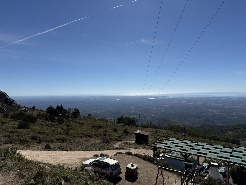 A bare hilltop with a stone hut, a shaded seating area and a parked car under overhead power cables, and a vast view over forested ridges down to the coast and the sea