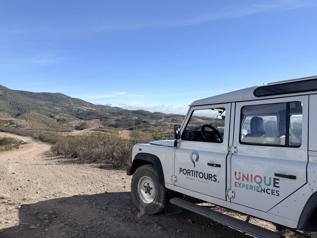 A white Land Rover Defender with PORTITOURS and Unique Experiences on its doors, parked on a gravel track among dry scrubby hills