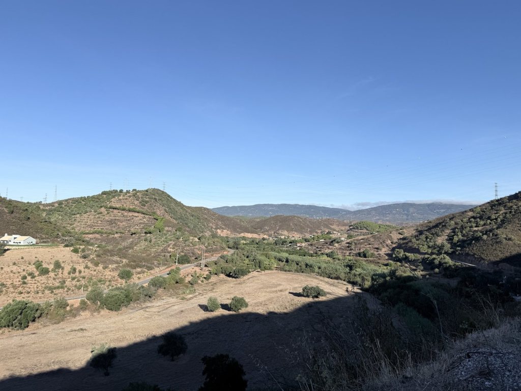 A valley of terraced hillsides and orchards with a road along the bottom, pylons along the ridges and cloud spilling over the mountains beyond