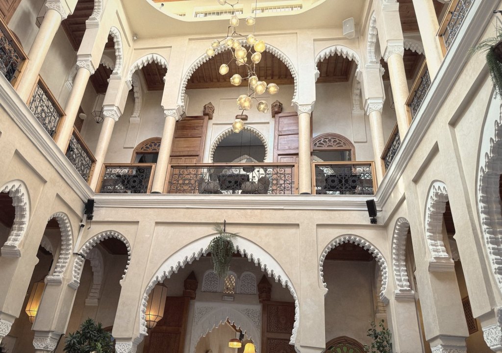 The courtyard of Dar Dada, looking up at two storeys of white scalloped arches and wooden balconies with iron railings, under a hanging chandelier of glass globes