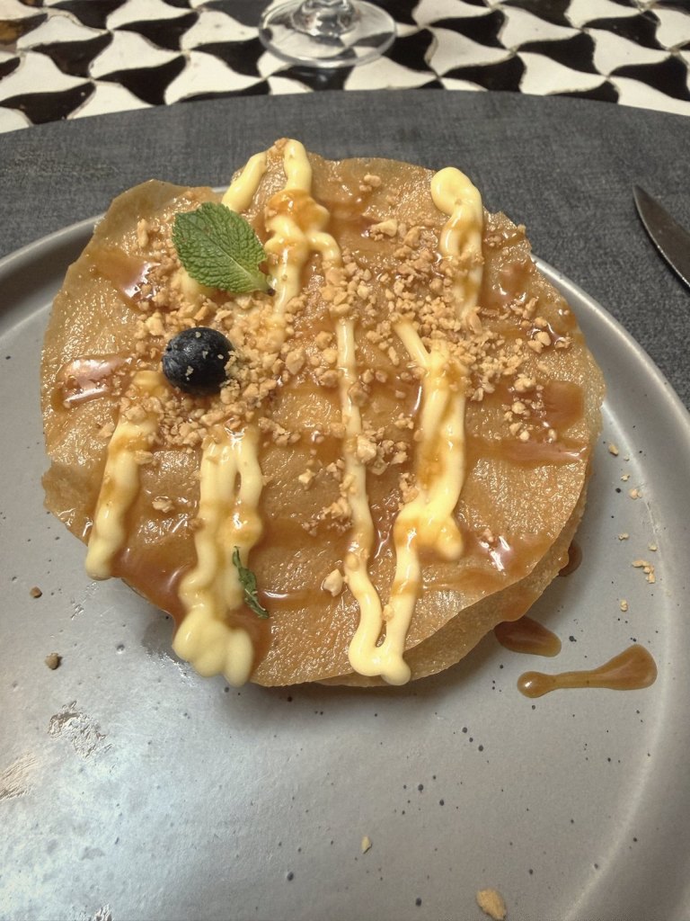 A round layered pastry dessert on a grey plate, drizzled with caramel and lines of custard and topped with crushed nuts, a mint leaf and a blueberry