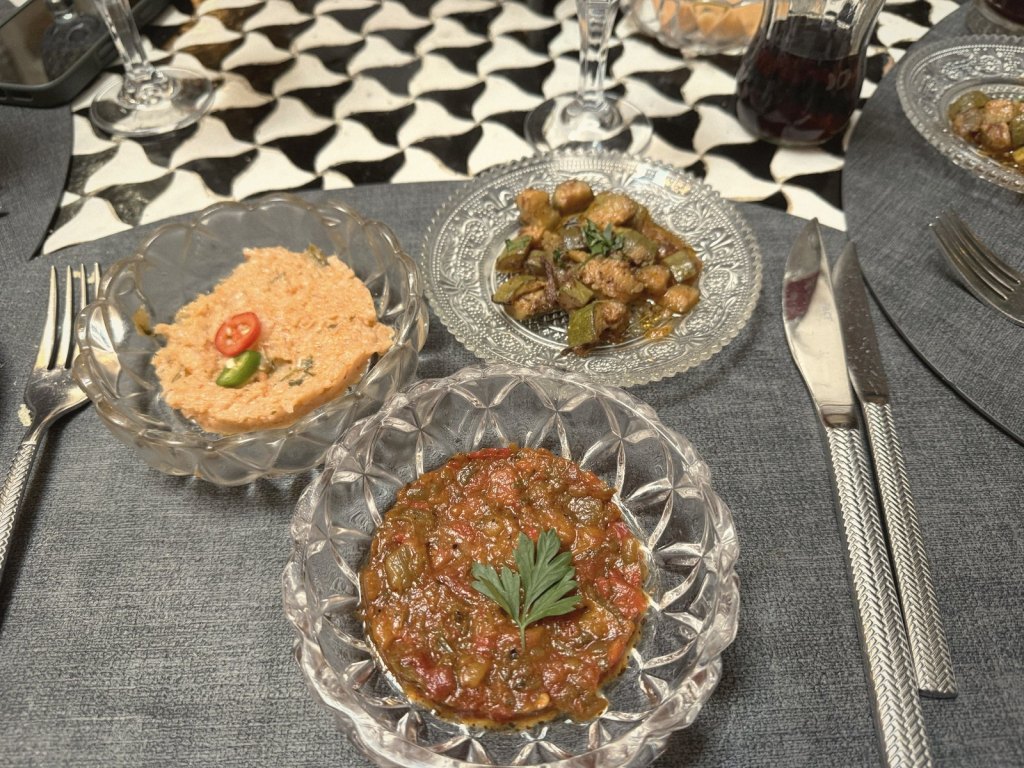 Three small Moroccan salads in cut-glass dishes on a grey place mat, a red cooked tomato and pepper salad, an orange spread topped with sliced chilli and a dish of courgette, with a black and white tiled table beyond