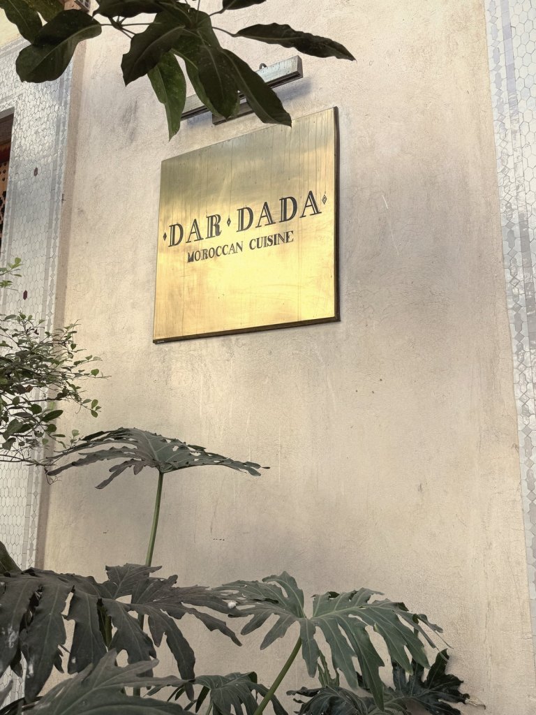 A brass plaque reading Dar Dada, Moroccan Cuisine, on a pale plastered wall edged with white tiles, with large green leaves in front