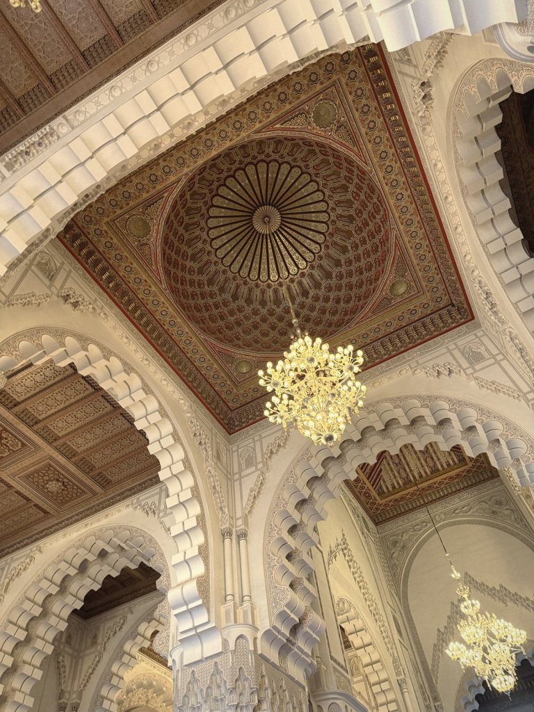 Looking straight up at a carved and painted wooden ceiling dome in red, gold and brown, with a lit chandelier hanging beneath it and tiers of white carved arches around it