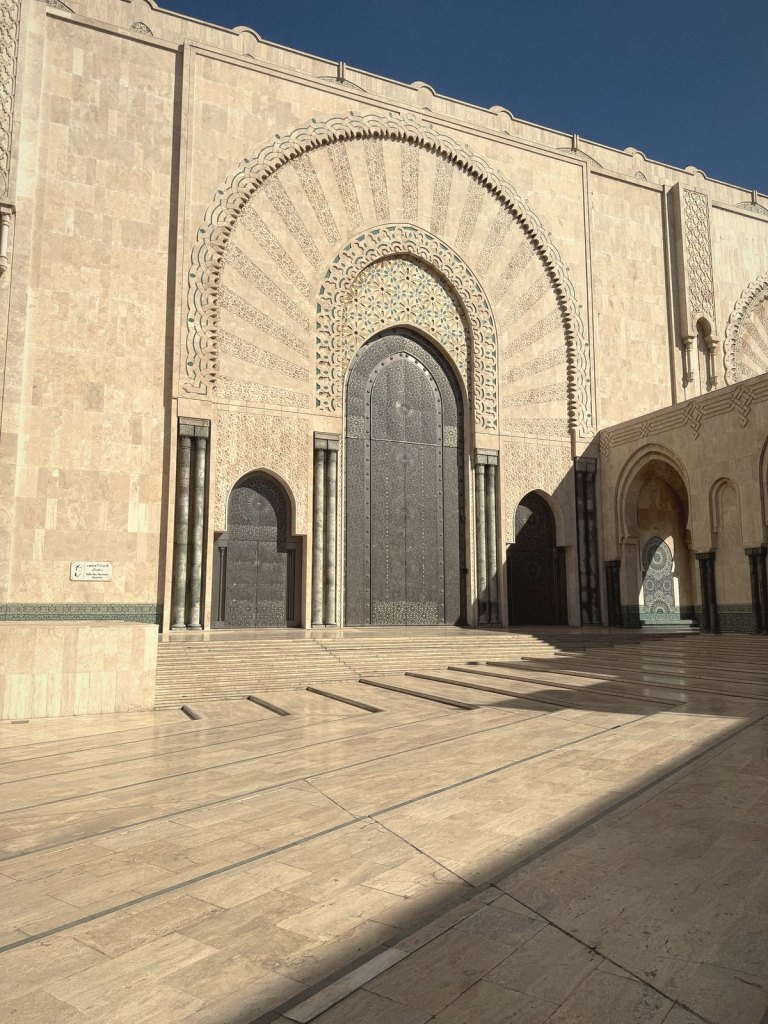 One of the mosque's entrances: a huge patterned metal door under a horseshoe arch of carved stone and tilework, with smaller doors either side and a polished stone courtyard in sun and shadow