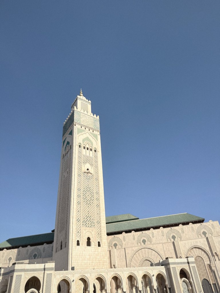 The minaret of the Hassan II Mosque against a deep blue sky, its pale stone carved in patterns, with a band of green and blue tilework near the top