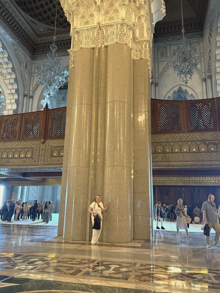 Jen standing at the foot of one of the prayer hall's giant polished stone pillars, which rises to a carved white capital, with carved wooden galleries behind and visitors crossing the marble floor