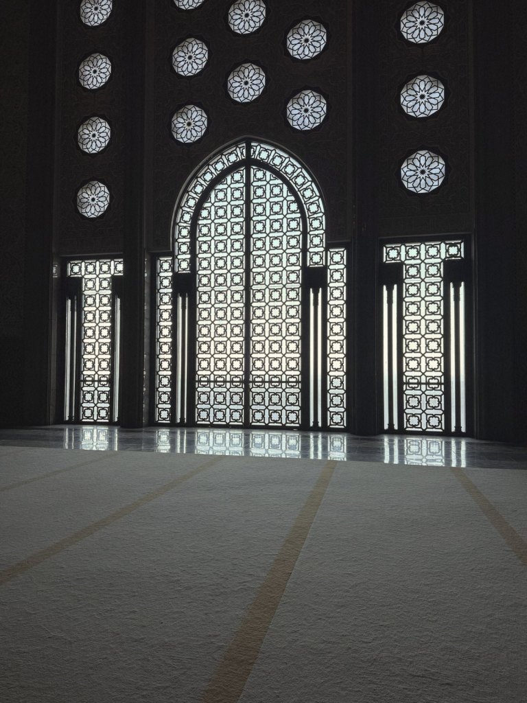 Tall lattice windows lit from outside along the dark wall of the prayer hall, with star-shaped openings above and a pale carpet running across the floor in front