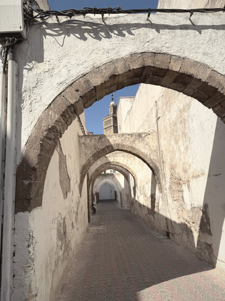 A narrow whitewashed lane running under a series of stone arches, with a minaret visible against the blue sky beyond