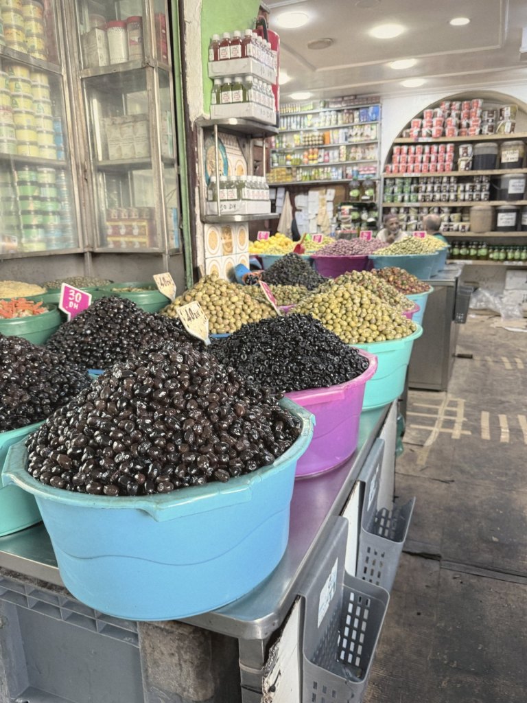 A shop front heaped with olives in turquoise and pink plastic tubs, black, green and red, with hand-written price cards, and shelves of jars and bottles behind