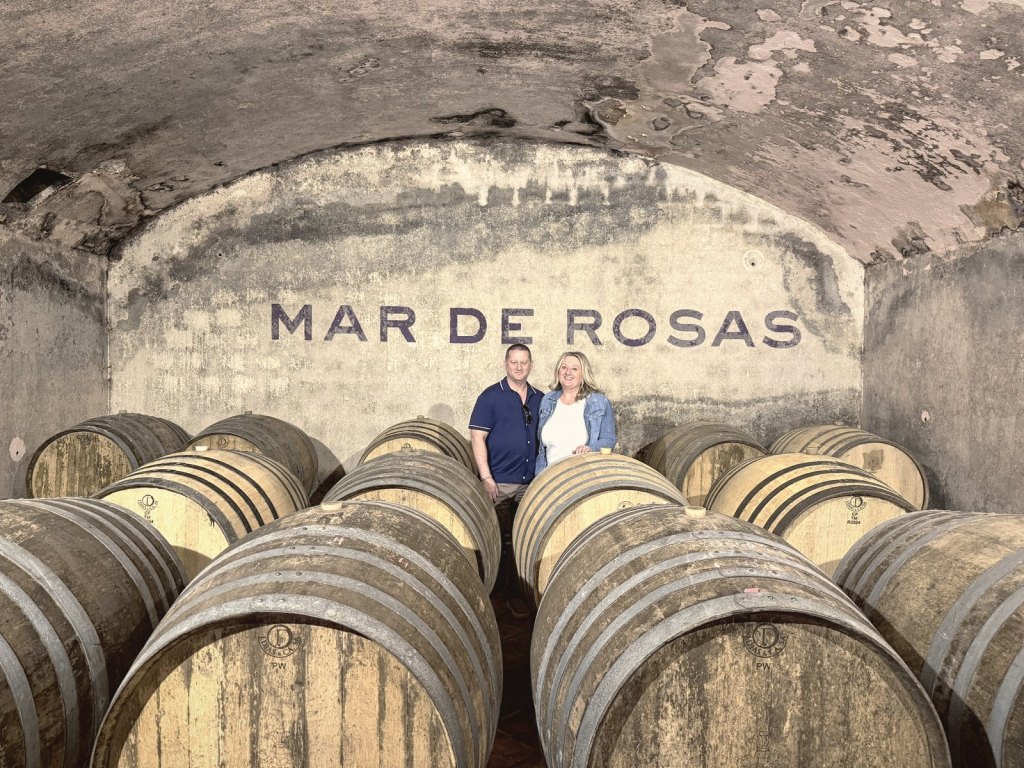 Chris and Jen standing between rows of barrels in front of MAR DE ROSAS painted in large letters across the end wall of the cellar