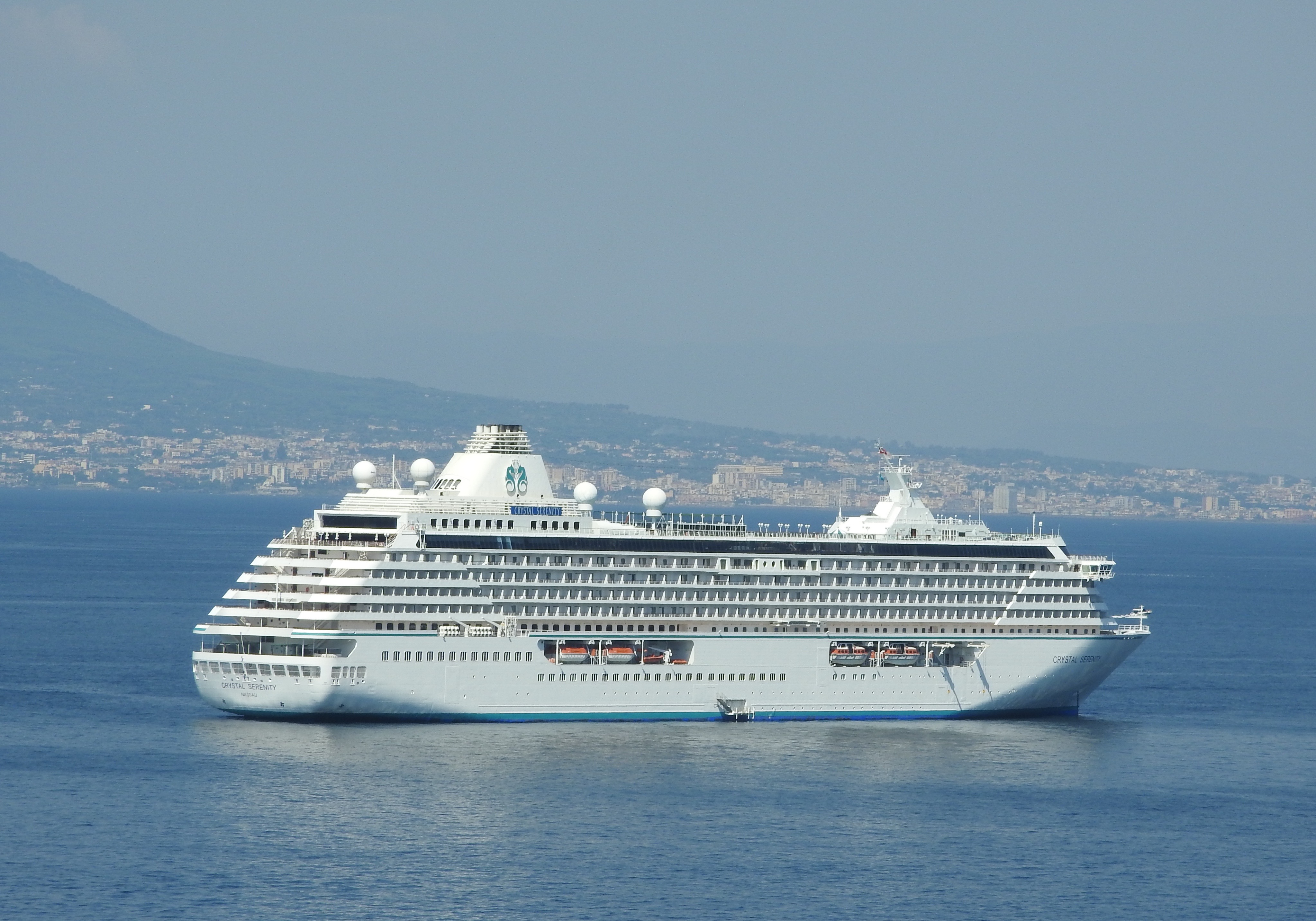 The Crystal Serenity at anchor in a calm blue bay, seen broadside, with a hazy mountain slope behind her and a town stretching along the far shore
