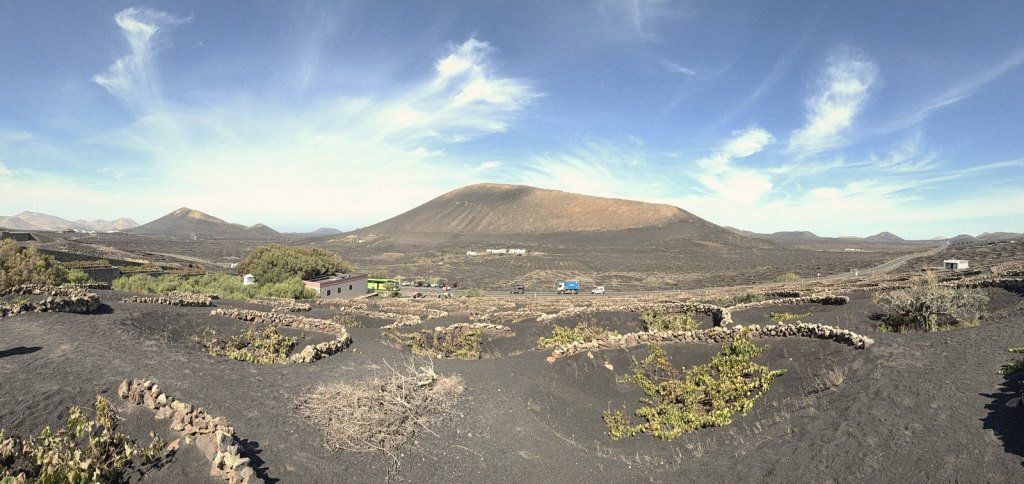 Panorama of La Geria: vines in walled pits of black ash, a road with cars and a bus, and a large volcanic cone under a blue sky.