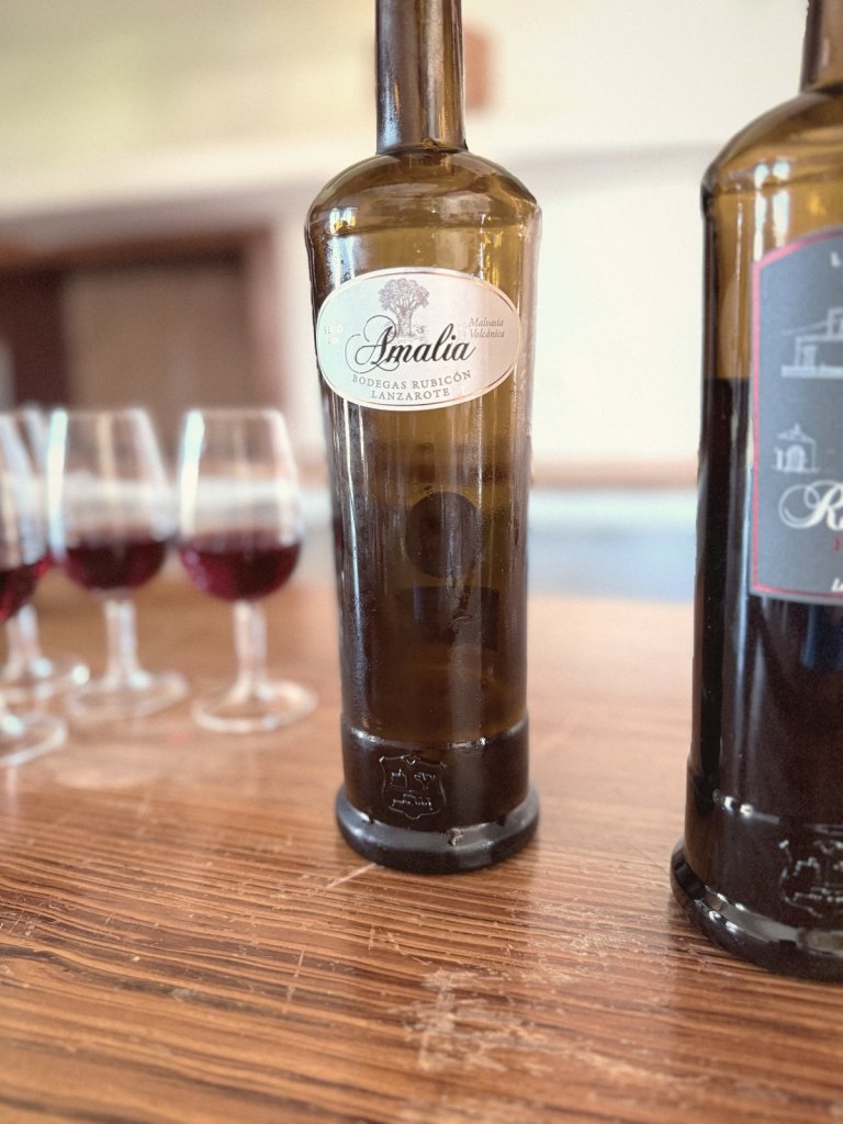 A bottle of Amalia dry Malvasía Volcánica from Bodegas Rubicón on a wooden counter, with glasses of red wine behind it.