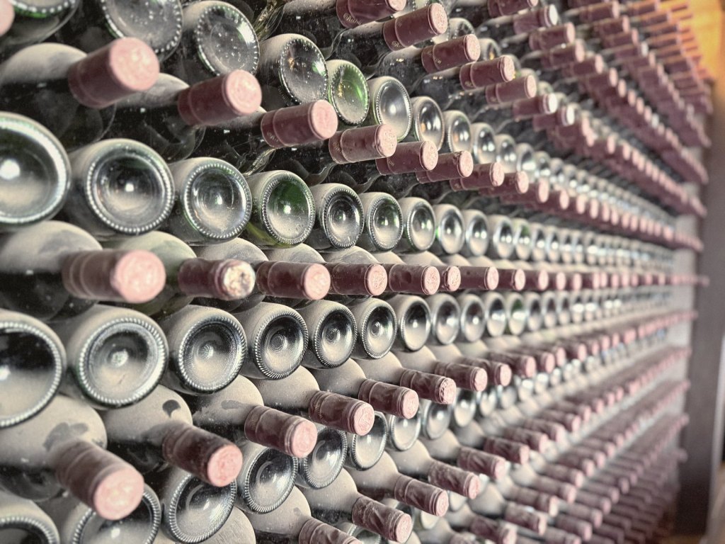 A wall of dusty wine bottles stacked on their sides, necks out, with red capsules.