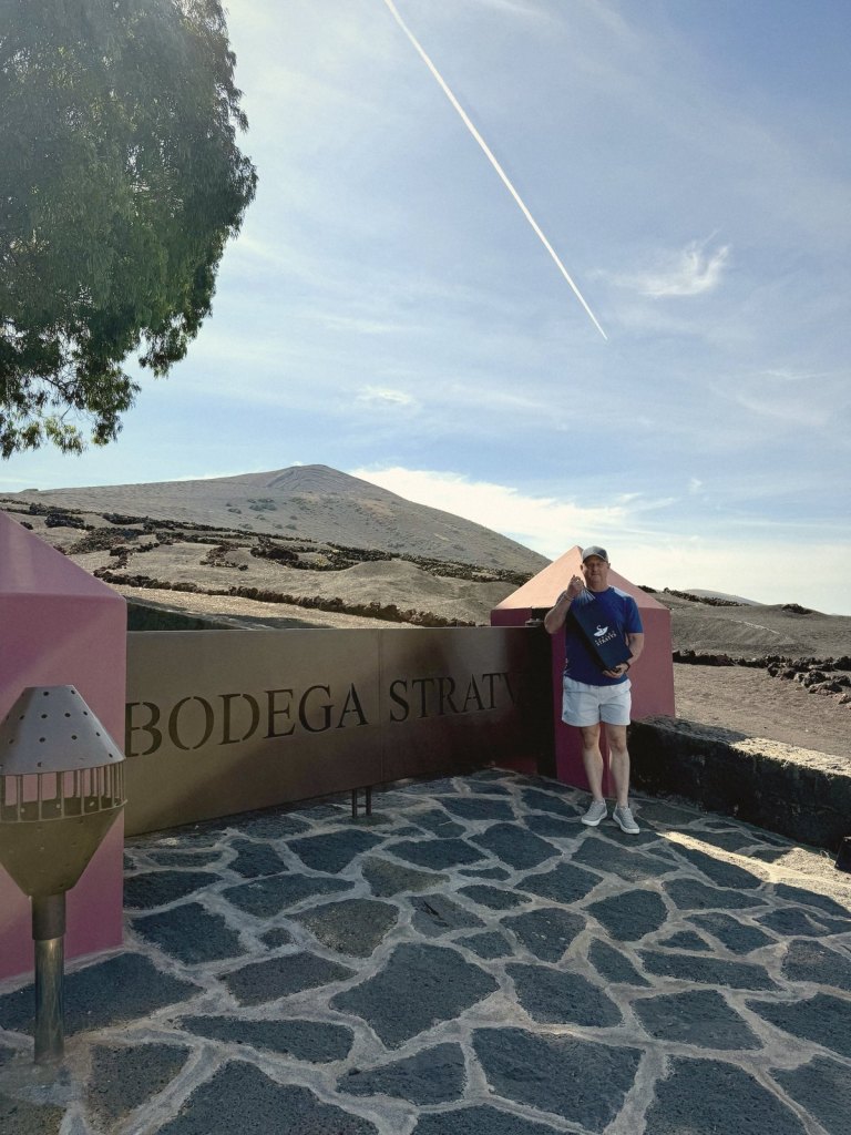 Chris standing by the Bodega Stratvs sign on a lava-stone terrace, holding a Stratvs bag, with walled vineyards and a volcanic cone behind.