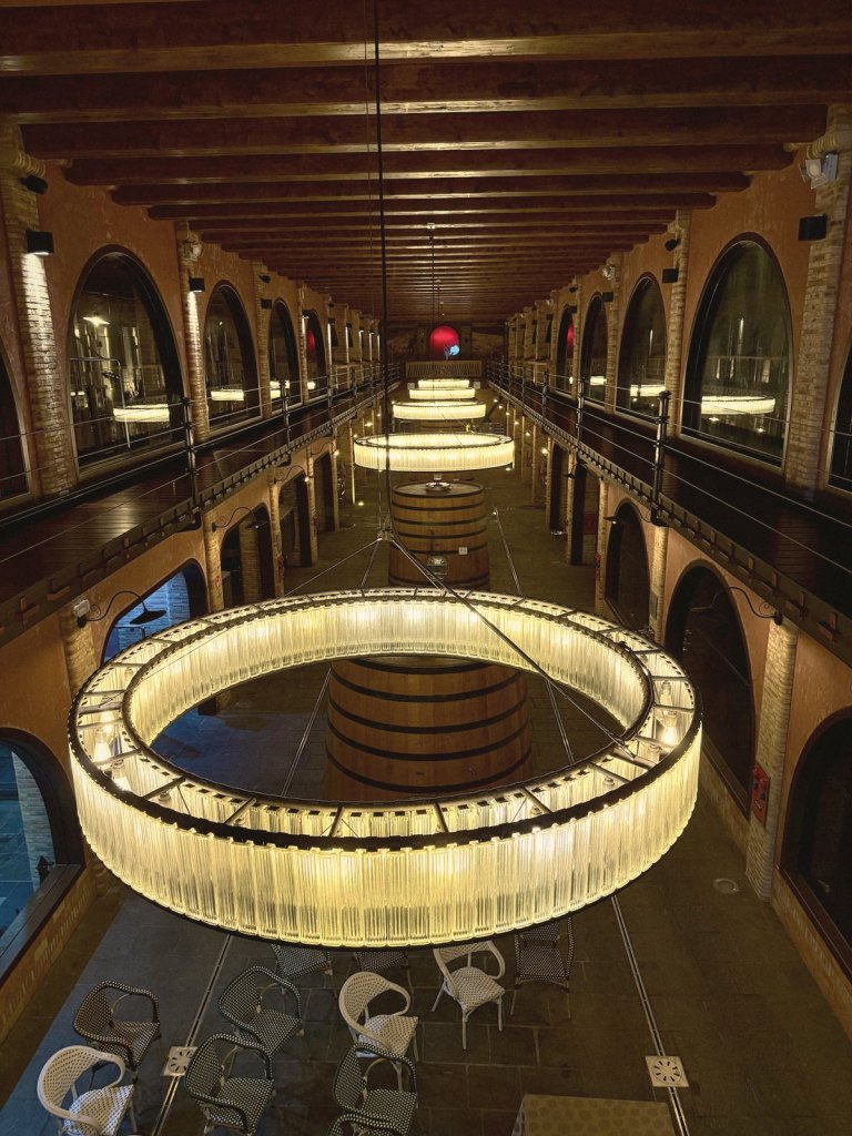 The Stratvs winery hall seen from the upper gallery: ring-shaped chandeliers, a giant wooden cask, and arched windows looking onto steel tanks.